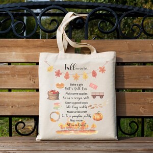 Peut inclure: Sac fourre-tout beige avec un motif sur le thème de l'automne. Le sac présente le texte "Fall to do list" avec des illustrations de feuilles, d'une tarte, de pommes, d'un chariot et d'une citrouille. Le sac a de longues anses.