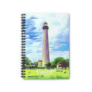 Op de afbeelding: Een spiraalgebonden notitieboek met een schilderachtige illustratie van een hoge vuurtoren tegen een blauwe lucht met wolken. Op de voorgrond is groen gras en kleine gebouwen te zien. De tekst Vintage Shore staat onderaan in het wit.