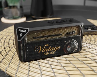 Puede incluir: Un altavoz Bluetooth rectangular de color negro con un panel frontal de malla dorada. El dispositivo cuenta con una pantalla de frecuencia estilo radio, una rejilla de altavoz circular a la derecha y el texto Vintage Shore en letras doradas.
