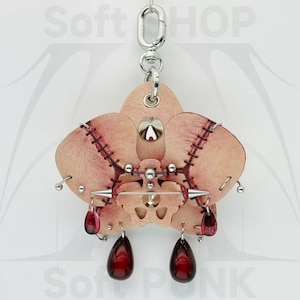 May include: A leather orchid-shaped keychain with a silver clasp. The light brown leather is stitched with red thread and adorned with silver studs and spikes. Two red teardrop-shaped charms dangle from the bottom. The text "Soft HOP" and "Soft PUNK" are visible in the background.