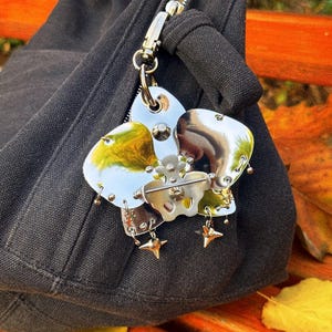 May include: A silver-toned metal flower-shaped charm with a pin, attached to a black bag. The charm has multiple petals with a reflective surface, accented with small star-shaped charms and dangling details. The bag is made of a dark fabric.