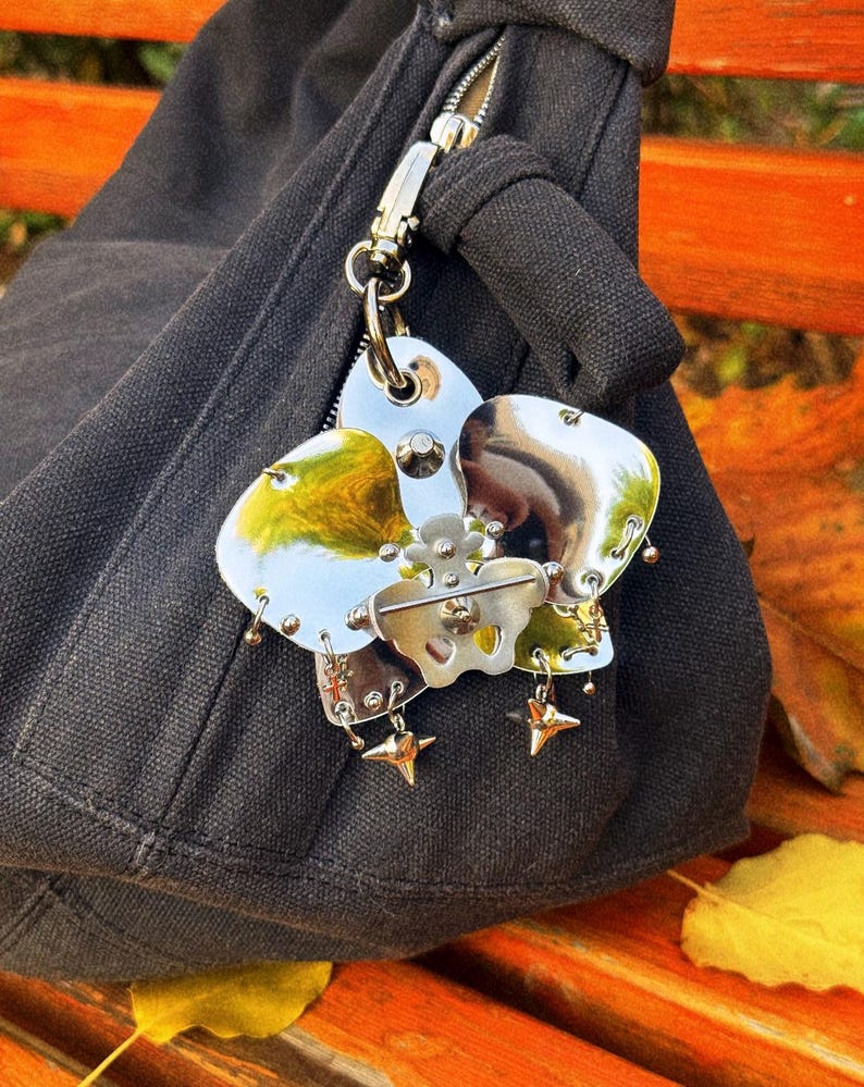 May include: A silver-toned metal flower-shaped charm with a pin, attached to a black bag. The charm has multiple petals with a reflective surface, accented with small star-shaped charms and dangling details. The bag is made of a dark fabric.