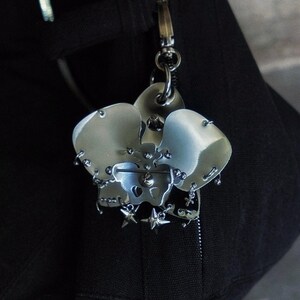 May include: A silver-toned metal keychain featuring a stylized skull design with star accents. The keychain is attached to a black fabric item with a zipper and metal hardware.