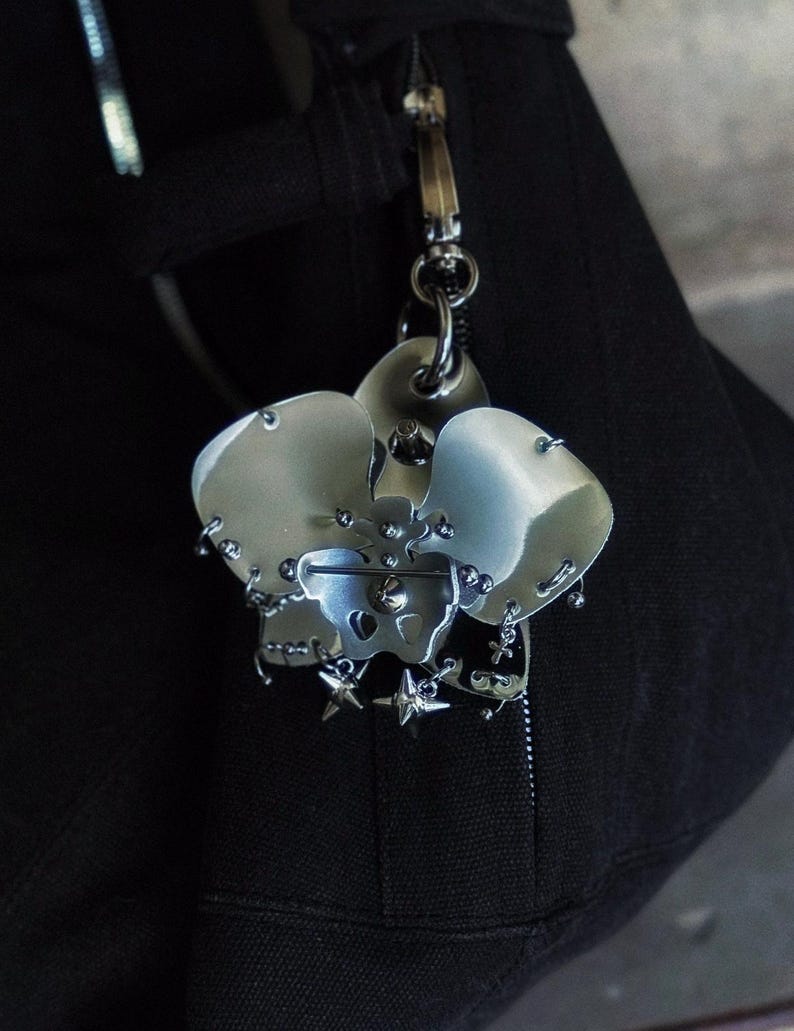 May include: A silver-toned metal keychain featuring a stylized skull design with star accents. The keychain is attached to a black fabric item with a zipper and metal hardware.