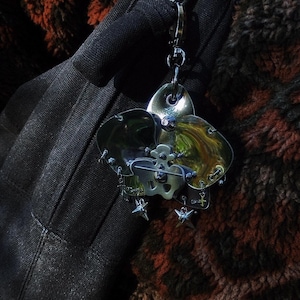 May include: A silver-toned metal brooch with a floral design, featuring three petal-shaped elements and a central skull motif. The brooch is adorned with small star and cross charms, attached to a chain. The background is a dark, textured fabric.