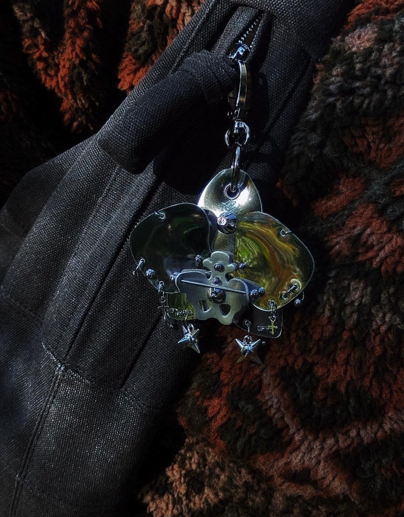 May include: A silver-toned metal brooch with a floral design, featuring three petal-shaped elements and a central skull motif. The brooch is adorned with small star and cross charms, attached to a chain. The background is a dark, textured fabric.