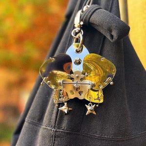 May include: A unique, metallic charm with a silver clasp, attached to a black fabric. The charm features a central silver piece with gold-toned accents and dangling star-shaped embellishments. The background is blurred with autumn colors.