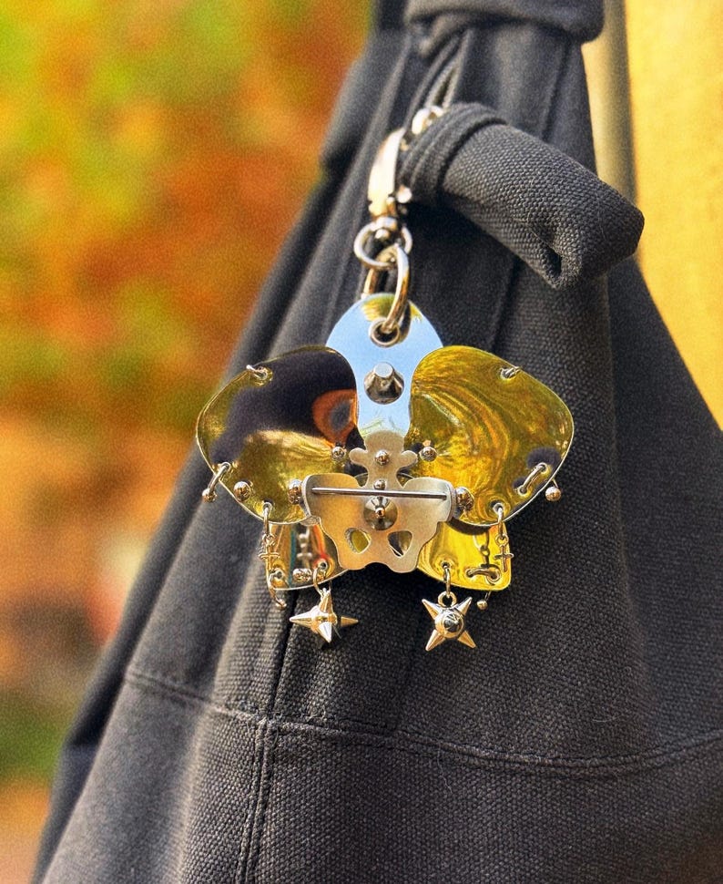May include: A unique, metallic charm with a silver clasp, attached to a black fabric. The charm features a central silver piece with gold-toned accents and dangling star-shaped embellishments. The background is blurred with autumn colors.