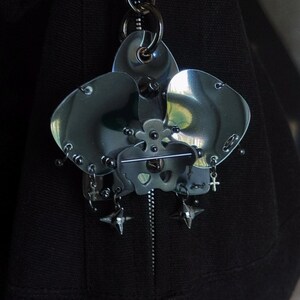 May include: A dark, metallic orchid-shaped keychain. The flower has three large petals and intricate details, including small rivets and dangling charms. The keychain is attached to a silver clasp and a black fabric background.