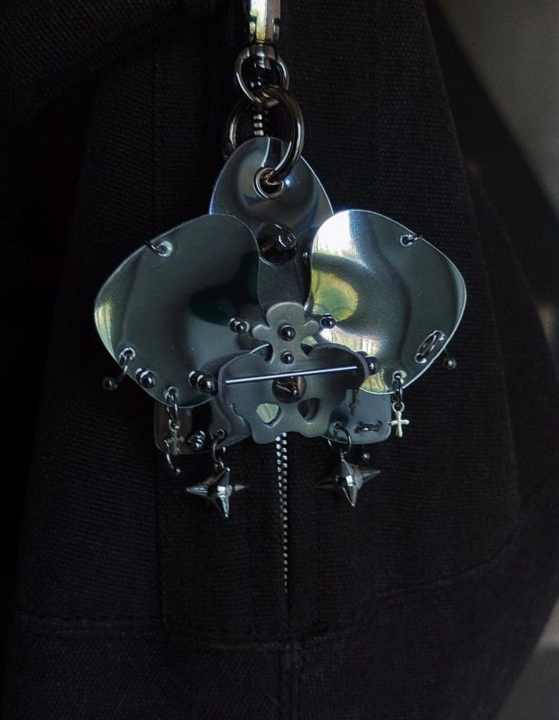 May include: A dark, metallic orchid-shaped keychain. The flower has three large petals and intricate details, including small rivets and dangling charms. The keychain is attached to a silver clasp and a black fabric background.