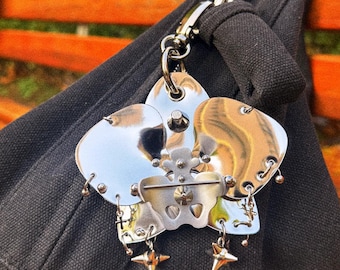 Könnte beinhalten: Eine silberfarbene Metallbrosche in Form einer Blume mit mehreren Blütenblättern und sternförmigen Anhängern, die von unten hängen. Die Brosche ist an einer schwarzen Stofftasche mit einem silbernen Verschluss befestigt.