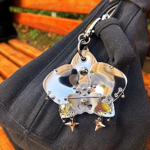 May include: A silver-toned metal keychain with a flower-like design, featuring multiple petals and star-shaped charms. The keychain is attached to a black fabric bag with a silver zipper and clasp. The background shows a wooden bench.
