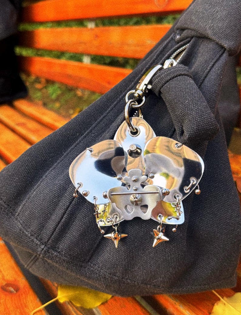 May include: A silver-toned metal keychain with a flower-like design, featuring multiple petals and star-shaped charms. The keychain is attached to a black fabric bag with a silver zipper and clasp. The background shows a wooden bench.