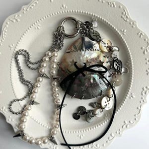 Puede incluir: Una colección de joyas y accesorios sobre un plato blanco decorativo. Los artículos incluyen un collar de perlas con puntas plateadas, una cadena de plata, un cordón negro y un colgante de concha adornado con botones y estrellas.