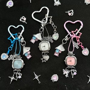 Puede incluir: Tres llaveros de reloj cuadrados con cierres en forma de corazón y detalles de estrella. Cada reloj tiene una esfera de diferente color: blanco, azul claro y rosa. Los dijes incluyen estrellas, cruces y adornos de gemas. Dispersos hay dijes adicionales de estrellas y gemas.