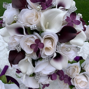 May include: A bridal bouquet with white roses, calla lilies, and purple flowers. The bouquet is decorated with silver and pearl embellishments.