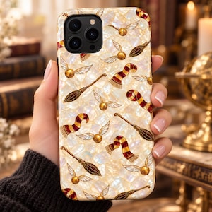 May include: A phone case with a pearlescent, iridescent white background featuring a repeating pattern of small golden winged balls, brown broomsticks, and red and yellow striped scarves. The case has a cutout for a triple-lens camera system.