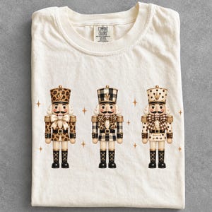 Può includere: Una t-shirt color crema con una grafica raffigurante tre schiaccianoci. Il primo ha un'uniforme con stampa leopardata, il secondo un motivo a quadri bianco e nero e il terzo un design a pois. Piccole icone a forma di stella dorata sono sparse.