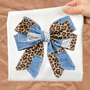 May include: A large decorative bow with a patchwork design. The bow features denim blue fabric, leopard print fabric, and white lace accents. The bow is on a white background.