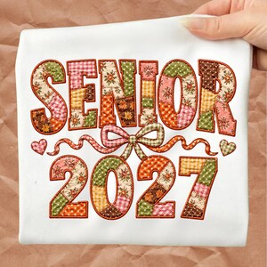 以下が含まれることがあります： パッチワークキルト風の刺繍が施された「SENIOR 2027」の文字が入った白いTシャツ。文字と数字は花柄、ギンガムチェック、水玉模様など様々なカラフルなパターンで埋め尽くされており、中央のリボンと2つのハートがアクセントになっています。