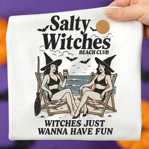 May include: White t-shirt with a graphic of two witches in beach chairs, the text "Salty Witches Beach Club" and "Witches Just Wanna Have Fun". The witches are wearing black hats and bikinis, with a broom and bats.