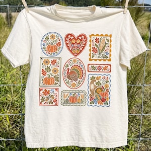 May include: A cream-colored short-sleeve t-shirt featuring a grid of nine colorful autumn-themed illustrations. The designs include a turkey, pumpkins, corn, acorns, and floral patterns in shades of orange, red, yellow, green, and blue.