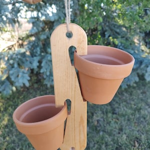 Op de afbeelding: Een hangende houten plantenbak met twee terracotta potten. Het houten frame is lichtbruin met een natuurlijke afwerking en de potten zijn warm, aards oranje. Een touwtje is bevestigd om op te hangen, ideaal voor kleine planten.