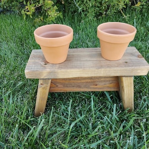 May include: Two terracotta flower pots sit on a small, rustic wooden bench. The bench has a natural wood finish and four legs. The pots are empty and have a classic, rounded shape. The scene is set outdoors on a bed of green grass.