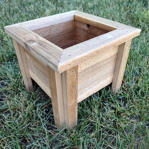 Può includere: Una fioriera quadrata in legno con finitura naturale. La fioriera ha un bordo superiore smussato ed è sostenuta da quattro robuste gambe. La fioriera è su erba verde, ideale per il giardinaggio.