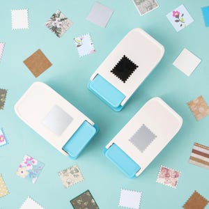 May include: Three white and blue stamp-shaped paper punches with a square stamp design. Scattered around are various patterned square paper cutouts in floral, geometric, and solid colors. The background is a light blue.