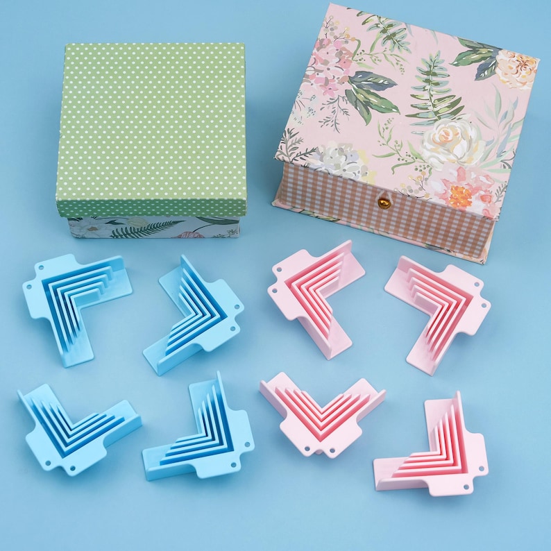 May include: An overhead shot shows two decorative boxes and several corner protectors. The boxes have floral and polka dot patterns. The corner protectors are light blue and pink. The background is a solid light blue.