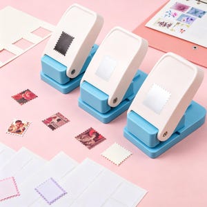 May include: Three blue and white stamp-shaped paper punches with a collection of colourful postage stamps and cut-out paper squares. The stamps feature floral and portrait designs. A pink notebook is visible in the background.