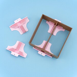 May include: Pink plastic corner braces and a brown cardboard box with dividers. The braces are in a T-shape and are designed to fit into the corners of the box. The background is a solid light blue.