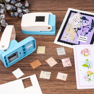 May include: A craft scene featuring two blue and white stamp-shaped paper punches, various decorative stamps, and handmade greeting cards with floral and butterfly designs. The stamps are in various colors and patterns.