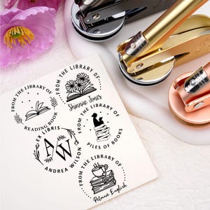 May include: An assortment of library seals in gold, copper, and black, alongside a white card featuring various circular library stamp designs. The stamps include floral and book illustrations, with text such as "From the Library of" and names.