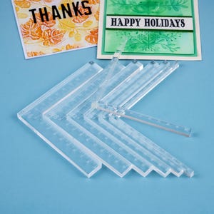 May include: Clear acrylic rulers and angle finders are arranged on a blue surface. Two greeting cards with the words "THANKS" and "HAPPY HOLIDAYS" are in the background. The rulers have inch markings.