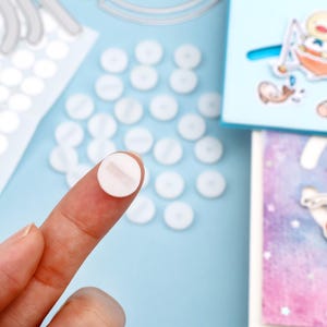 Op de afbeelding: Een close-up van een vinger die een kleine, witte, ronde zelfklevende stip vasthoudt met een witte streep in het midden. Verschillende vergelijkbare stippen zijn verspreid over een lichtblauwe ondergrond, samen met een stansvel en een kleine kaart met een cartoonillustratie.