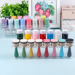 Op de afbeelding: Een verzameling cosmetische producten, waaronder kleine flesjes met witte doppen en make-up applicators met gekleurde handvatten. De items zijn gerangschikt in doorzichtige plastic display stands. Een ingelijste aardbeienillustratie staat op de achtergrond.