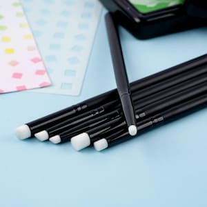 Puede incluir: Un conjunto de brochas de maquillaje negras con cerdas blancas, dispuestas sobre una superficie azul claro. Las brochas varían en tamaño y forma, con algunas plantillas en el fondo. Las brochas están limpias y parecen sin usar.