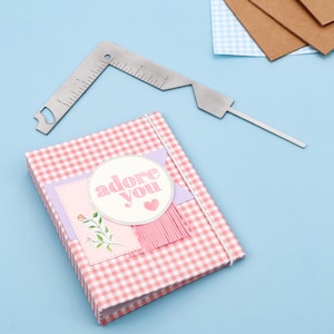 以下が含まれることがあります： 「adore you」と書かれた装飾ラベルとピンクのタッセルが付いた、ピンクと白のギンガムチェック柄のジャーナル。銀色の金属製の定規がジャーナルの上に配置されています。背景には追加の文房具があります。