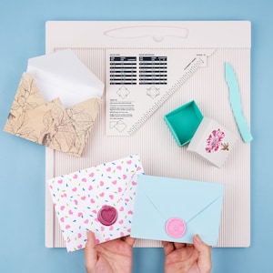 May include: An assortment of stationery supplies, including envelopes, a scoring board, a box, and a craft knife. Two envelopes are sealed with pink wax seals. One envelope has a floral design, and the other has a pattern of pink hearts.