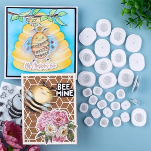 May include: A selection of craft items with bee motifs. The image shows two cards with bee illustrations and text, alongside white, circular embellishments. One card reads "Oh! Christmas Bee" and the other says "BEE MINE".
