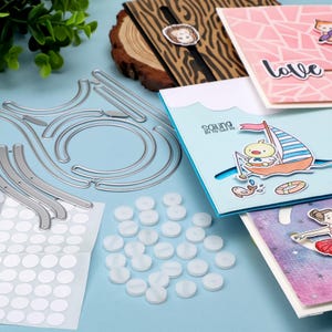 May include: A selection of craft supplies including metal die cuts, white adhesive dots, and several handmade greeting cards. One card features a sailboat illustration with the text "Sailing by to say hi."