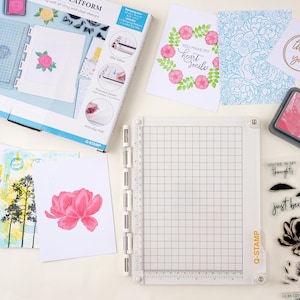 May include: A white Q-STAMP stamping platform with a grid pattern, surrounded by stamped floral designs on white cards. The cards feature pink and blue floral patterns, and the text "YOU MAKE MY heart smile". A box with the text "Q-STAMP" is also visible.