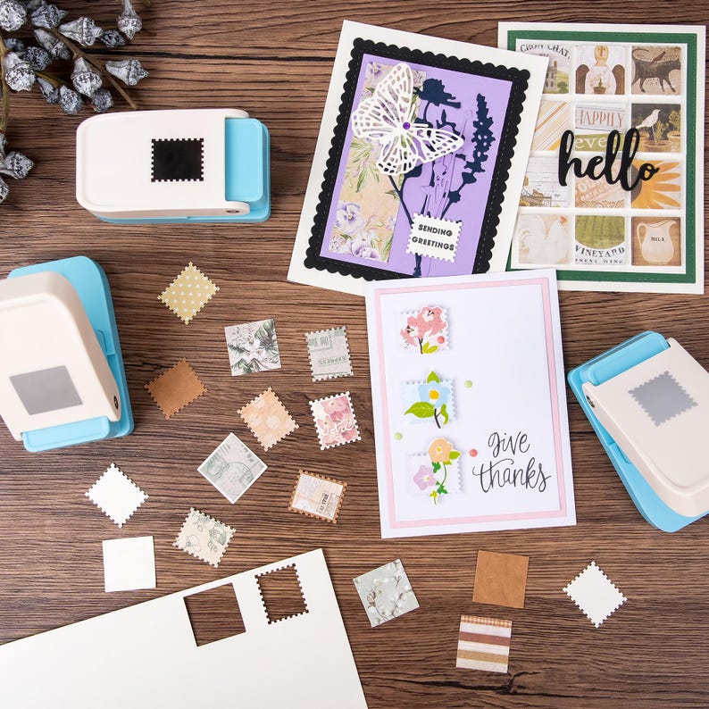 May include: A craft scene with handmade greeting cards and stamp-shaped paper cutouts. The cards feature floral designs, butterfly cutouts, and the words "hello" and "give thanks." Two blue and white stamp-shaped paper punches are also visible.