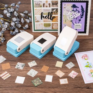 May include: Three blue and white stamp-shaped paper punches with various cut-out stamps and cards on a wooden surface. The cards have floral and butterfly designs. One card says "hello".