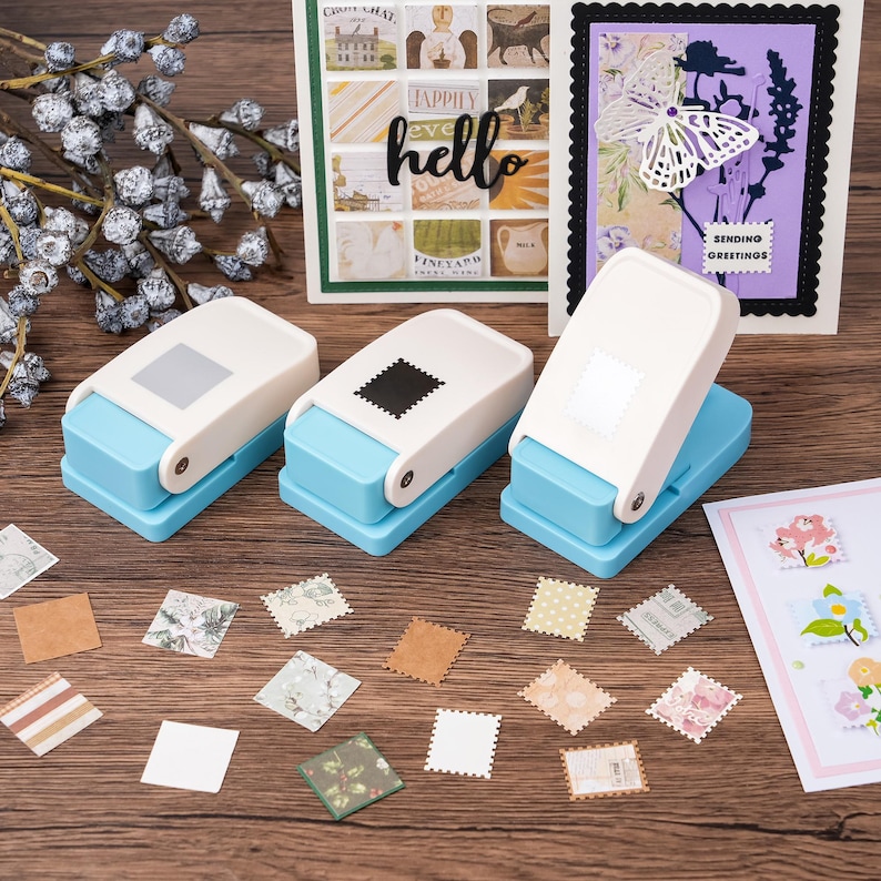 May include: Three blue and white stamp-shaped paper punches with various cut-out stamps and cards on a wooden surface. The cards have floral and butterfly designs. One card says "hello".