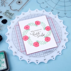 May include: A white card with a floral wreath design and the text "YOU MAKE MY heart smile" is centered on a white, circular craft tool with degree markings. Additional stamps and ink pads are visible.