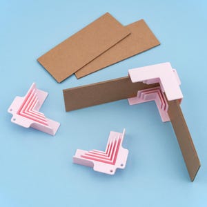May include: Pink corner protectors holding brown cardboard pieces, with two additional cardboard pieces on a blue background. The corner protectors are designed to fit the cardboard pieces. The image shows a crafting or packaging theme.