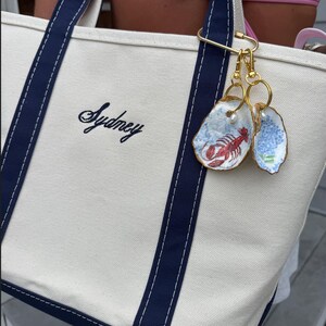 May include: A cream-colored canvas tote bag with navy blue trim and handles, featuring the embroidered name Sydney in black script. Attached to the handle is a gold-toned metal clip holding two oyster shell charms, one painted with a red lobster and one with blue flowers.